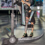Apocalyptic scythe-wielder cosplay by @sayun_kc at Anime NYC 2025 Javits Center, featuring gray wig, tactical black outfit with holographic panels, tattoos, and massive mechanical scythe – animenyc.com.