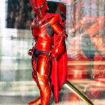 Solo Leveling crimson knight cosplay by @sirjuliuscosplay at Anime NYC 2025 Javits Center, featuring full red armor, horned helmet, flowing cape, golden sword, and dramatic mirrored aura effect – animenyc.com.
