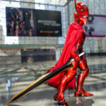 Solo Leveling crimson knight cosplay by @sirjuliuscosplay at Anime NYC 2025 Javits Center, featuring full red armor, horned helmet, flowing cape, golden sword, and dramatic mirrored aura effect – animenyc.com.