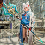 Feixiao cosplay from Honkai: Star Rail by @smugycos at Anime NYC 2025 Javits Center, featuring silver wig, fox ears, blue embroidered tunic, white coat, and ornate teal-and-gold polearm – animenyc.com.