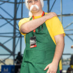 Sakamoto Days cosplay at Anime NYC 2025 Javits Center featuring Taro Sakamoto with yellow shirt, green apron, glasses, and white mustache – animenyc.com.