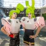 @rinn.tuu and @shio.kou wearing flamboyant cosplay with green sculpted wigs, jewel-embellished white armor tops, and pink fringe sleeves at Anime NYC 2025 Javits Center – animenyc.com.