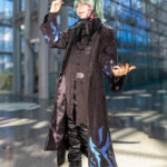 @otomia.a cosplaying Baby from the Saja Boys at Anime NYC 2025 in Javits Center, wearing a black coat with purple and blue flames, mint green wig, tall hat, and platform boots – animenyc.com.