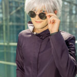 @astra.tuu cosplaying Satoru Gojo from Jujutsu Kaisen at Anime NYC 2025 in Javits Center, wearing a dark high-collared jacket, white spiky wig, and black sunglasses – animenyc.com.
