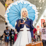 @lizwoop cosplaying Violet Evergarden with blue parasol, white dress, brown gloves, emerald brooch, and suitcase at Anime NYC 2025 inside Javits Center – animenyc.com.