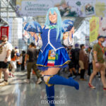 @emi.st0ner_bang cosplaying Aqua from Konosuba in a blue dress, thigh-high boots, aqua wig, and holding folding fans while posing playfully at Anime NYC 2025 inside Javits Center – animenyc.com.