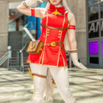 @nicopokie cosplaying Gold Ship from Uma Musume: Pretty Derby in red and gold outfit with horse ears, tail, and salute pose at Anime NYC 2025 Javits Center – animenyc.com.