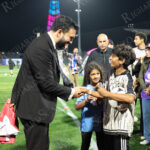 Zohran Mamdani in a black suit and tie bends down to shake hands with two smiling children on a soccer field during “The Cost of Living Classic” in Coney Island.