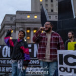 Two protest organizers speak into microphones in front of a large banner reading “ICE OUT OF NYC – STOP THE DEPORTATIONS” during the ICE OUT of NYC protest at Foley Square on October 22, 2025.