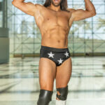 Cosplayer @themachodoc poses as Macho Man Randy Savage at New York Comic Con 2025, flexing his muscles while wearing black trunks with white stars, white boots, a “MADNESS” bandana, and signature sunglasses.