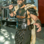 Cosplayer @justanotherhero dressed as a Viking warrior poses with a raised axe at New York Comic Con 2025, wearing black leather armor, fur shoulder guards, and war paint under bright Javits Center lighting.