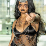 Cosplayer @surprimity dressed as the Enchantress from Suicide Squad poses at New York Comic Con 2025, wearing a crescent moon headpiece, black lace outfit with chains, and rune markings on her face.