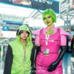 Cosplayers @shea.briar and @deviilbby dressed as GIR and Zim from Invader Zim pose together at New York Comic Con 2025, wearing green and pink costumes with bright makeup and wigs inside the Javits Center.