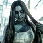 Cosplayer @barbiechulanyc dressed as a ghostly horror spirit at New York Comic Con 2025, with cracked gray body paint, white eyes, long black hair, and a distressed white gown inside the Javits Center.