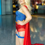 Cosplayer @kailcarps dressed as Supergirl poses at New York Comic Con 2025, wearing a blue rhinestone corset, red glitter shorts, red cape, and platform boots with blonde hair and a confident smile.