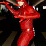 Cosplayer @calebweekss dressed as Daredevil from Marvel Comics poses in a combat stance at New York Comic Con 2025, wearing a red armored suit and helmet with horns while holding red batons.