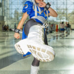 Cosplayer @barythaya dressed as Chun-Li from Street Fighter performs a high kick at New York Comic Con 2025, wearing a blue and gold dress, white boots, and spiked wristbands inside the Javits Center.