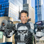 Cosplayer @heroes_4_hire_costumes dressed as The Punisher at New York Comic Con 2025, wearing black tactical armor with a white skull emblem and holding two prop guns in a battle stance outdoors in NYC.