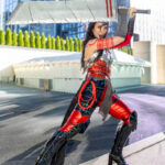Cosplayer @cos.wonder dressed as Red Sonja poses in a battle stance outside New York Comic Con 2025, holding a large sword overhead while wearing metallic red and silver armor with red markings and high boots.