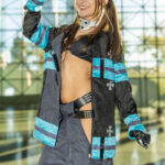 Cosplayer @racksonrays dressed as Tamaki Kotatsu from Fire Force poses at New York Comic Con 2025, flexing her arm confidently in a firefighter-style outfit with blue reflective stripes and a black bikini top inside the Javits Center.