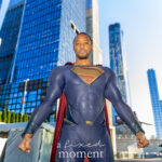 Cosplayer @supermazing_23 dressed as Superman poses powerfully in a blue and red suit with a cape amid New York City skyscrapers at New York Comic Con 2025.