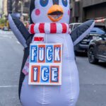 A person in an inflatable penguin costume with a red-and-white scarf stands on a New York City street holding a protest sign reading “FUCK ICE” during the No Kings March on October 18, 2025.