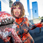 Cosplayer @bombshellbeckie dressed as a Venom/Carnage hybrid snarls and poses with a large symbiote claw prop and fanged mouth under bright daylight outside the Javits Center during New York Comic Con 2025.