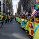 A large group of protesters marches through Midtown Manhattan holding a bright yellow “No Kings” banner and waving American flags during the No Kings March on October 18, 2025, in New York City.