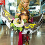 Cosplayer @lumi_veneri dressed as a Night Elf warrior from World of Warcraft poses inside the Javits Center during New York Comic Con 2025, holding a large dual-bladed glaive decorated with leaves and glowing designs.