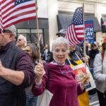 An older woman wearing a burgundy outfit smiles and waves an American flag during the No Kings March in New York City on October 18, 2025, surrounded by fellow demonstrators holding flags and banners.