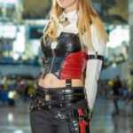 Cosplayer @clockychan dressed as a modern Harley Quinn poses inside the Javits Center during New York Comic Con 2025, wearing a red and black leather corset, white shirt, and tactical pants with utility straps.