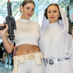 Cosplayers @bohemiansongstress and @meljp93 dressed as Princess Leia from Star Wars pose together at New York Comic Con 2025, one in a white action outfit with a blaster and the other in a white gown with a hood and silver belt.
