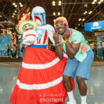 Cosplayers @floris_lilium and @nanaakaonasis pose at New York Comic Con 2025. One wears Mandalorian armor styled with Puerto Rican colors and a flowing red and white skirt while holding a Grogu doll, and the other wears a tropical outfit with a mint-green shirt, denim shorts, and yellow sunglasses.