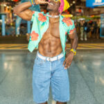 Cosplayer @nanaakaonasis poses at New York Comic Con 2025 wearing a mint-green Hawaiian shirt with orange flowers, denim shorts, a yellow beanie, pink wig, yellow sunglasses, and white sneakers, striking a confident pose with one finger pointing upward.