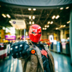 Cosplayer @ryfam_ dressed as Red Hood points a prop gun toward the camera at New York Comic Con 2025, wearing a red armored helmet, brown trench coat, and tactical gear with a red bat symbol on the chest under the Comic Con banner.