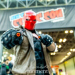 Cosplayer @ryfam_ dressed as Red Hood aims two prop pistols at the camera beneath the New York Comic Con banner, wearing a red helmet, brown jacket, and black armor with a red bat symbol.
