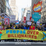 A large crowd of protesters marches through Midtown Manhattan holding a long yellow banner that reads “NO KINGS NEW YORK” and “PEOPLE OVER BILLIONAIRES,” surrounded by colorful signs and puppets during the No Kings March in New York City on October 18, 2025.