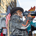 A protester wearing a black hat holds a megaphone and shouts during the No Kings March in New York City on October 18, 2025, with an American flag on his back and protest signs visible in the background.
