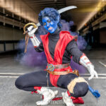 Cosplayer @toxiekat dressed as Nightcrawler from X-Men at New York Comic Con 2025, crouching in a dynamic pose with a sword and purple smoke behind them, wearing a red and black outfit with blue skin and a tail at the Javits Center in NYC.