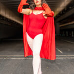 Cosplayer @kenndragonn dressed as Scarlet Witch from Marvel Comics at New York Comic Con 2025, posing with hands raised in a red leotard, cape, gloves, and headpiece at the Javits Center in NYC.