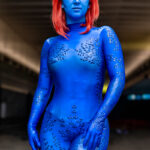 A cosplayer dressed as Mystique from X-Men at New York Comic Con 2025, covered in blue body paint and textured scales with red hair and yellow eyes, standing confidently at the Javits Center in NYC. @kenndragonn
