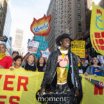 A smiling protester wearing a black hat walks beside others holding a yellow banner and a large “Liberty Not Tyranny” torch sign during the No Kings March in New York City on October 18, 2025.