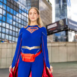 Cosplayer @samcosplayys dressed as Supergirl at New York Comic Con 2025, wearing a blue bodysuit with the red and gold “S” emblem, red boots, cape, and fanny pack, posing confidently outside the Javits Center in NYC.