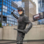 Cosplayer @matthew_dion20 dressed as Daredevil from Marvel Comics at New York Comic Con 2025, posing with batons in a tactical black suit with red highlights and a horned mask outside the Javits Center in NYC.
