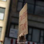 A hand holds up a cardboard protest sign that reads “IMPEACH TRUMP (again)” during the No Kings March in New York City on October 18, 2025.