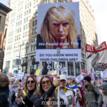 A protester wearing sunglasses holds a sign that reads “We Found Aunt Tifa” and “Do You Know Where Your Children Are?” during the No Kings March in New York City on October 18, 2025, surrounded by other demonstrators holding banners and flags.