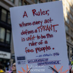 A protest sign at the No Kings March in NYC reads “A Ruler, whose every act defines a TYRANT, is unfit to be the ruler of a free people – as stated in 1776, Declaration of Independence,” held high among city buildings and fellow protesters.