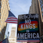 A colorful protest sign reading “We the People – No Kings in America” is held aloft with American flags waving nearby amid tall buildings during the No Kings March in New York City on October 18, 2025.