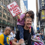 A smiling man carries his young daughter on his shoulders as she holds a red-and-white protest sign that reads “No King Any Time” during the No Kings March in New York City on October 18, 2025.