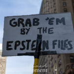 A handmade protest sign reading “Grab ’Em by the Epstein Files” held above the crowd during the No Kings March in New York City on October 18, 2025, with tall Midtown buildings in the background.